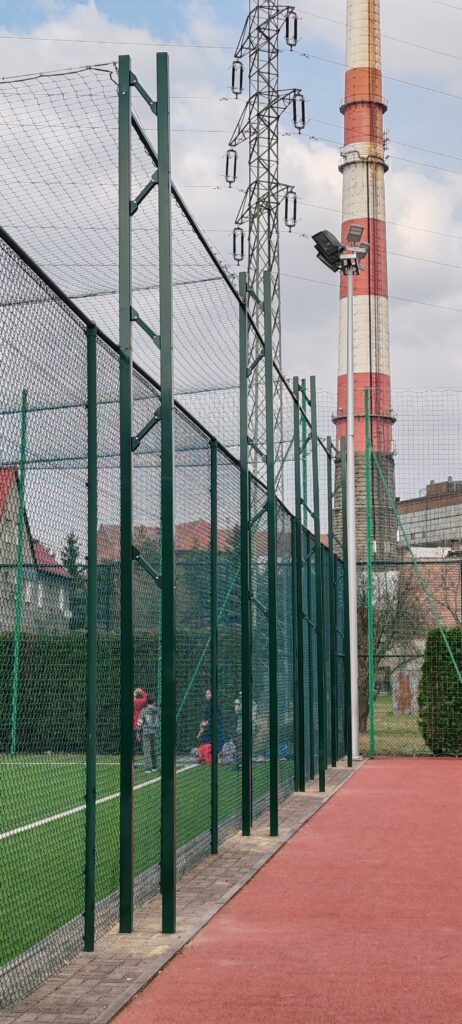 Boisko z siatką ochronną, lampy boiska, linie energetyczne i komin przemysłowy. Konstrukcja zadaszenia ze wspornikami. realizacja siatki ochronnej